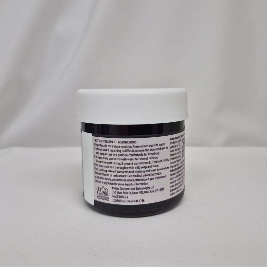 A small plastic jar of black liquid with a white screw cap, showing the back label with first-aid instructions, set against a draped white fabric background.