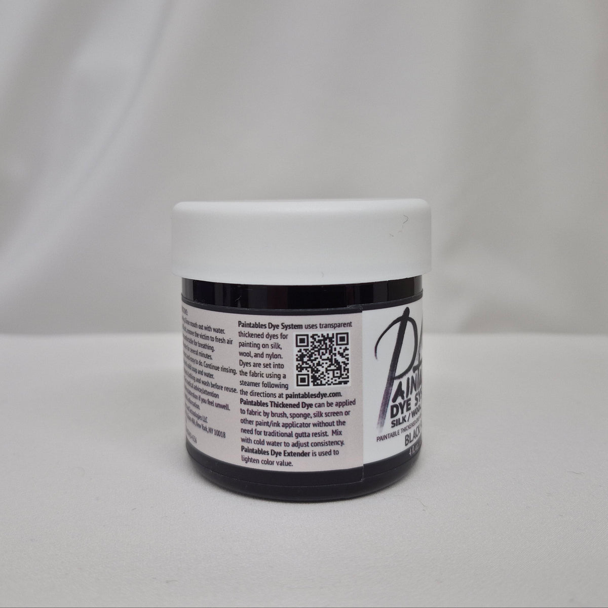 A small plastic jar of black liquid with a white screw cap, showing the side label with information on Paintables Dye System, set against a draped white fabric background.