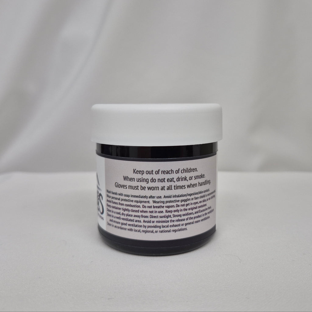 A small plastic jar of black liquid with a white screw cap, showing the back label with safety instructions, set against a draped white fabric background.