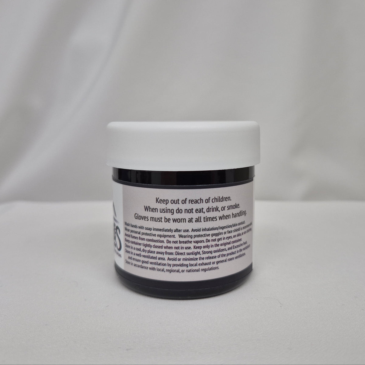 A small plastic jar of black liquid with a white screw cap, showing the back label with safety instructions, set against a draped white fabric background.