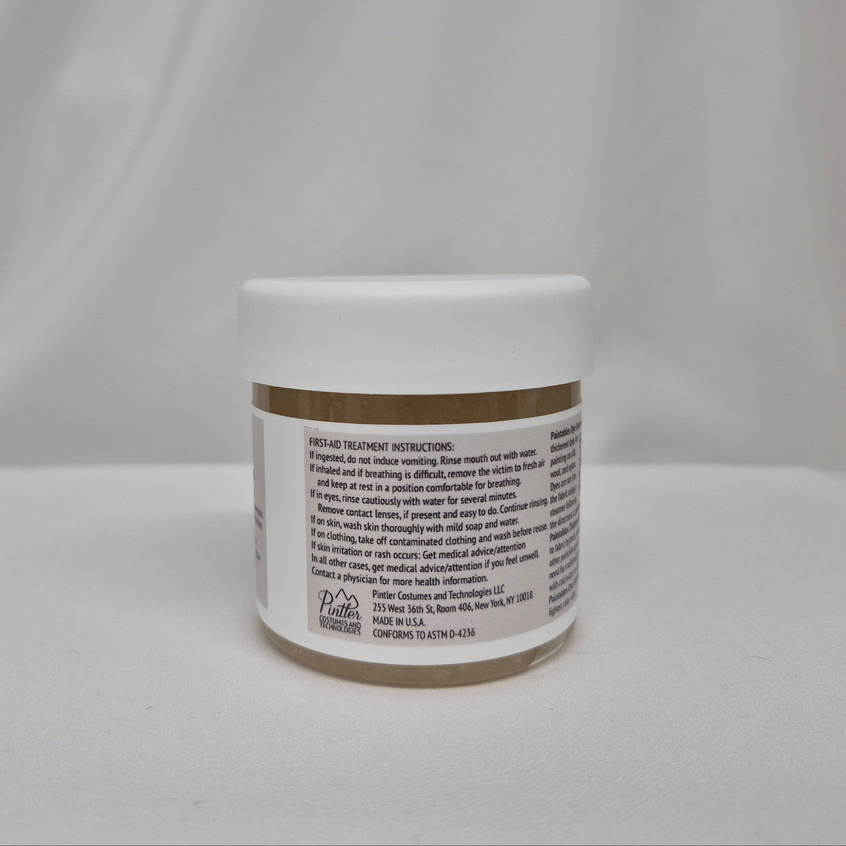 A small white plastic jar with a gum extender thickener inside, a white lid, and black label featuring black text containing first-aid instructions, set against a draped white fabric background.