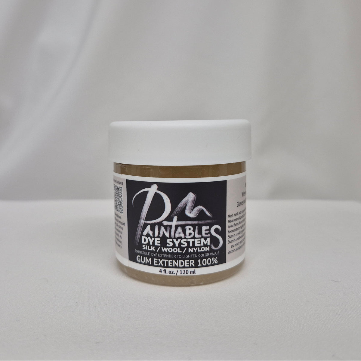 A small white plastic jar of Paintable Dye System Gum Extender 100% on a white fabric background.