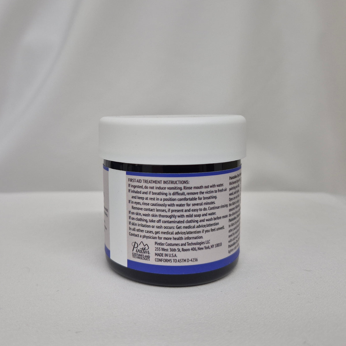 A small plastic jar of blue liquid with a white screw cap, showing the back label with safety instructions, set against a draped white fabric background.