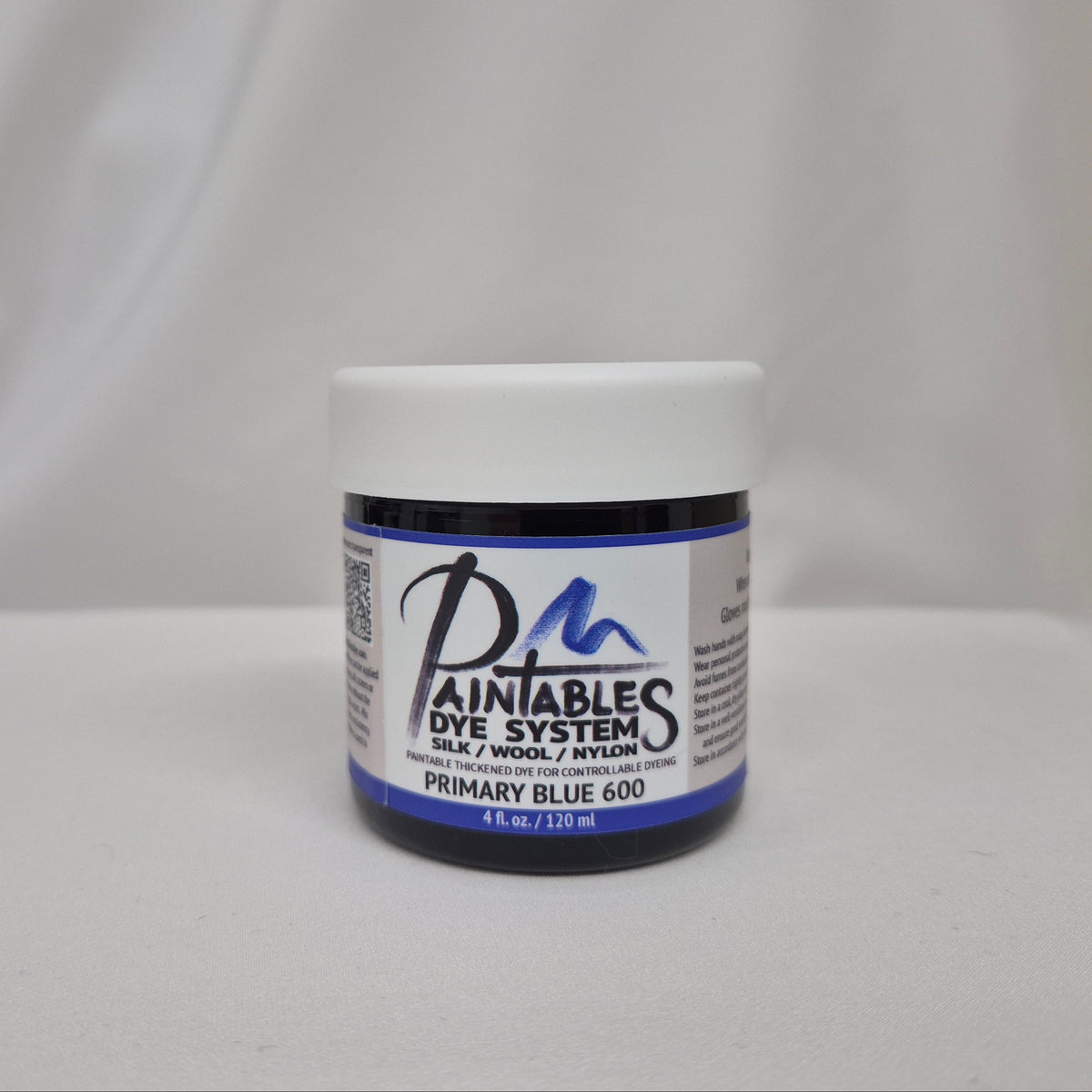 A jar of Paintables Dye System Primary Blue 600 dye, featuring a white lid and a white label with blue and black text, set against a draped white fabric background.