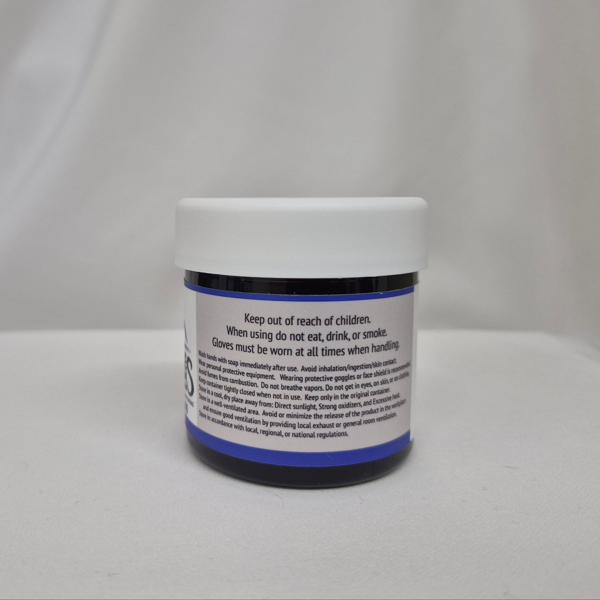 A small plastic jar of yellow liquid with a white screw cap, showing the back label with safety instructions, set against a draped white fabric background.
