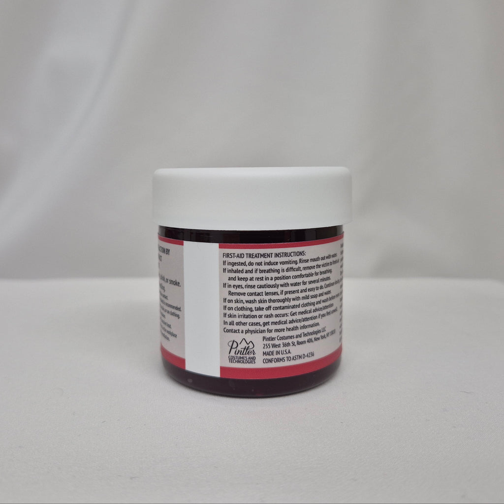 A small plastic jar of red liquid with a white screw cap, showing the back label with first-aid instructions, set against a draped white fabric background.