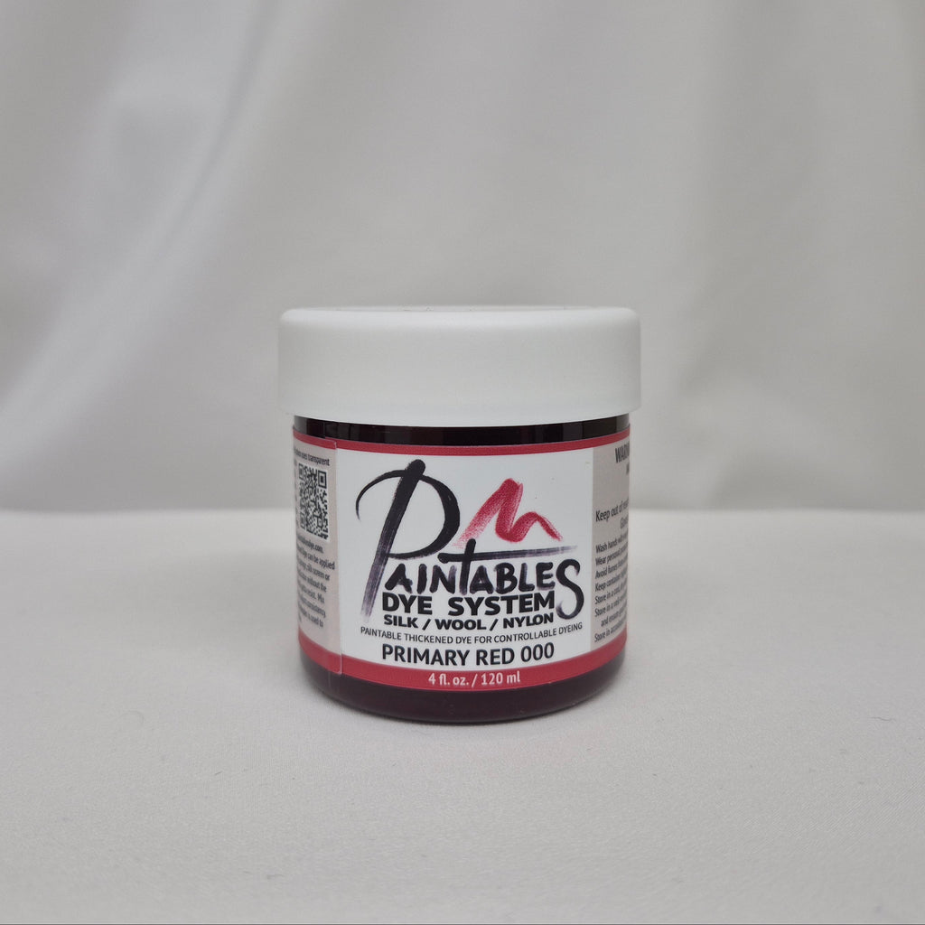 A small jar of Paintables Dye System Primary Red 000 dye, featuring a white lid and a white label with red and black text, set against a draped white fabric background.