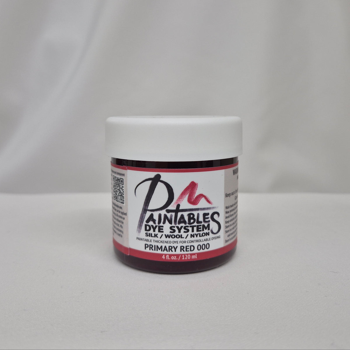 A small jar of Paintables Dye System Primary Red 000 dye, featuring a white lid and a white label with red and black text, set against a draped white fabric background.