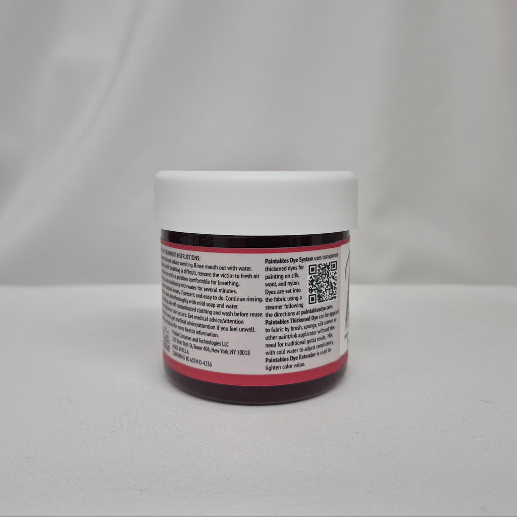A small plastic jar of red liquid with a white screw cap, showing the side label with information about Paintables Dye System, set against a draped white fabric background.