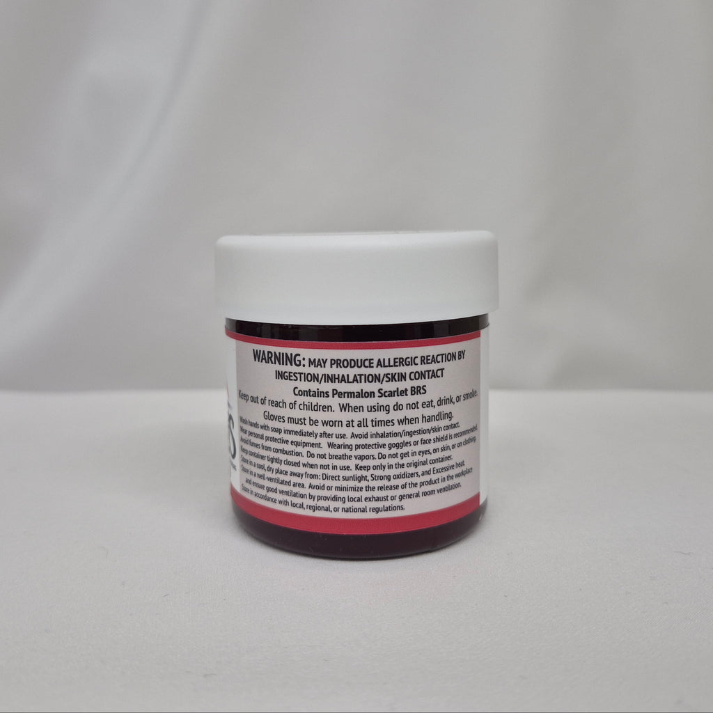 A small plastic jar of red liquid with a white screw cap, showing the back label with warning and safety instructions, set against a draped white fabric background.