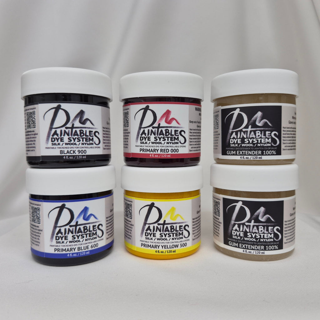 Six jars of Paintables Dye System dyes in Black, Blue, Red, Yellow and Extender, arranged in two rows of three against a draped white fabric background.