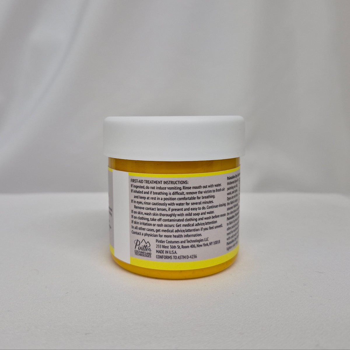 A small plastic jar of yellow liquid with a white screw cap, showing the back label with first-aid instructions, set against a draped white fabric background.