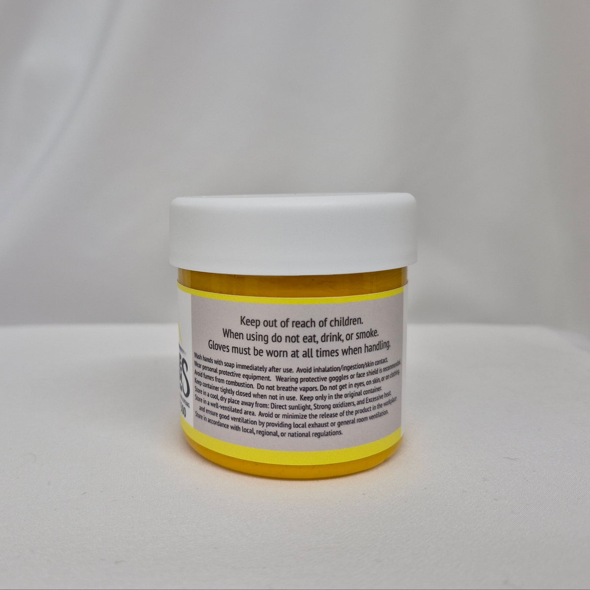 A small plastic jar of yellow liquid with a white screw cap, showing the back label with safety instructions, set against a draped white fabric background.